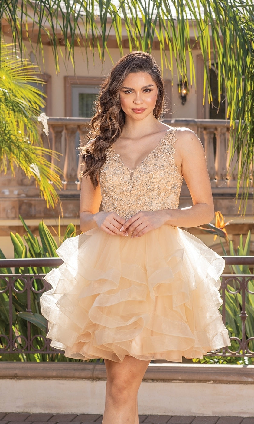 A woman stands outdoors in a Beaded-Bodice Tiered Short Homecoming Dress 3273, featuring a gold lace bodice and ruffled tulle skirt, with greenery and a balcony visible in the background.